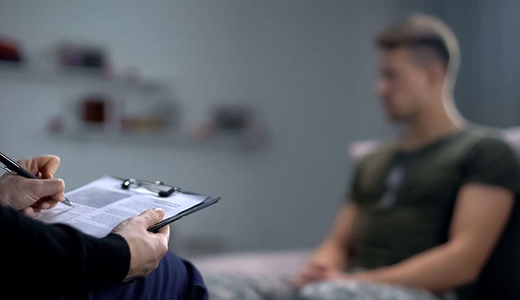 Psychologist making notes during therapy session with sad male soldier, PTSD